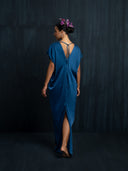 A woman with flowers in her hair stands barefoot against a dark background, wearing the Rimmba Gaya Silk Kaftan Dress in indigo—crafted from sustainable silk with a back slit and tie details.