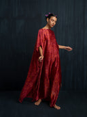 A barefoot woman in the Bellas Red Silk Kaftan - Eco Print by Rimmba | Sustainable Clothing stands gracefully against a dark background, purple flowers in her hair, eyes closed, and arms gently outstretched.