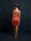A woman stands barefoot facing a dark wall, wearing the Mira Eco-Print Dress - Mini by Rimmba | Sustainable Clothing in hand-dyed red and orange shades, with a low back. Her hair is styled up and decorated with purple flowers.