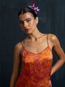 A woman stands before a dark background, calmly gazing at the camera in the Mira Eco-Print Dress - Mini by Rimmba | Sustainable Clothing. Her slicked-back hair is adorned with a purple flower.