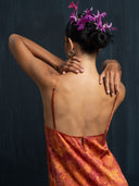 A person with short dark hair and purple flowers faces away, wearing the Mira Eco-Print Dress - Mini by Rimmba | Sustainable Clothing. One hand rests on their neck, the other arm is bent, set against a dark background.