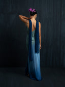 A woman faces away, wearing the Scarlette Silk Dress - Indigo Ombre by Rimmba | Sustainable Clothing and a blue scarf. Her hair is in a bun with purple flowers, left arm raised to her head, set against a dark background.