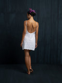 A woman stands barefoot with her back to the camera, wearing the Karima Dress—an embroidered white cotton mini dress by Rimmba | Sustainable Clothing—her hair in a bun with purple flowers, set against a dark textured background.