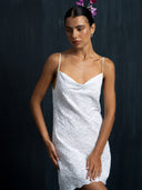 A woman in the Rimmba Karima Dress, a white cotton embroidered mini dress with thin straps, stands against a dark background. Pink flowers decorate her hair as she gazes downward, one hand softly resting on her thigh.