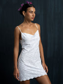 A woman in the Karima Dress - Cotton Embroidery by Rimmba | Sustainable Clothing stands against a dark backdrop, gazing to the side with flowers in her hair and small hoop earrings.