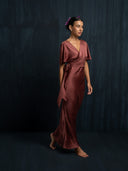 A woman with a flower headband wears the Ria Silk Dress in burgundy by Rimmba | Sustainable Clothing, standing barefoot against a dark textured background.