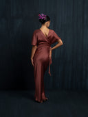 A woman stands with her back to the camera, wearing the Ria Silk Dress in burgundy by Rimmba | Sustainable Clothing, and a pink flower in her hair against a dark background, one hand on her hip, head turned slightly to the side.