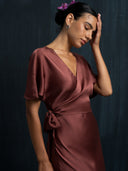 A woman in the Ria Silk Dress - Burgundy by Rimmba | Sustainable Clothing stands against a dark backdrop, hair up with purple flowers, gently touching her forehead and tilting her head forward.