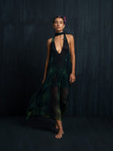 A woman stands barefoot against a dark backdrop, wearing Rimmba's Aphrodite Dress – Aurora: a flowing, sheer black and green adjustable silk dress with deep V-neckline, choker collar, open back, flowers in her hair, and a calm expression.
