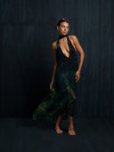 A woman stands barefoot against a dark background in the Aphrodite Dress - Aurora by Rimmba | Sustainable Clothing, featuring green and black patterns. With flowers in her hair and eyes closed, she poses gracefully, showcasing the open back design.