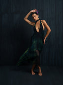 A barefoot woman poses gracefully against a dark background, wearing the Rimmba Aphrodite Dress - Aurora. With a deep neckline, flowing black and green fabric, and open back design, the look is completed by flowers in her hair and an arched arm above her head.