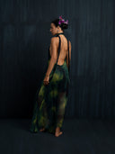 A woman with flowers in her hair stands barefoot on a dark floor, wearing the Rimmba Aphrodite Dress - Aurora. The adjustable silk dress with green and black patterns flows elegantly as she faces sideways against a dark backdrop.