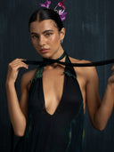 A person with slicked-back dark hair and pink flowers poses confidently in the Rimmba Aphrodite Dress - Aurora, holding the adjustable silk straps of this sustainable black dress against a dark textured background.