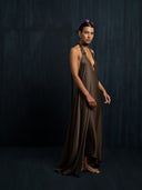 A woman with short hair stands barefoot against a dark textured background, wearing the Rimmba Aphrodite Dress - Iron Mud. The elegant halter-neck dress features a flowing brown design. She wears a crown decorated with small purple butterflies.
