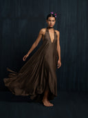 A woman with short dark hair and a serious expression stands barefoot against a dark backdrop, wearing the Rimmba Aphrodite Dress - Iron Mud. Two purple butterfly hairpins accent her look as the dramatic dress flares with her movement.