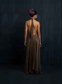 A woman with a bun and purple flowers stands facing away, wearing Rimmba's Aphrodite Dress - Iron Mud—a flowing backless gown—set against a dark, textured background.