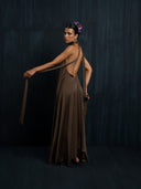 A woman stands barefoot against a dark background, wearing the flowing, backless Rimmba Aphrodite Dress - Iron Mud with long silk ribbons. Her hair is in a bun adorned with purple flowers as she looks over her shoulder at the camera.