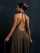 A woman faces away from the camera in Rimmba's Aphrodite Dress - Iron Mud, holding silk strap ribbons. Her dark bun is adorned with purple flowers, set against a dark textured background. Sustainable elegance by Rimmba.