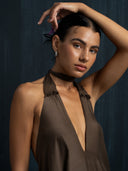 A woman with slicked-back dark hair and dewy skin poses in the Aphrodite Dress - Iron Mud by Rimmba | Sustainable Clothing, featuring a deep v-neck and sleeveless design, one arm raised against a dark textured background.
