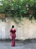 A woman in a Rimmba Ria Silk Dress - Burgundy stands barefoot on a stone path, facing a weathered wall draped with green vines and pink flowers. The sustainable dress is made from luxurious mulberry silk tinted with botanical dye.