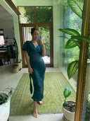A woman wearing the Rimmba Ria Silk Dress - Indigo takes a mirror selfie barefoot in a bright, plant-filled room. Sunlight streaming through large windows highlights the sustainable Mulberry Silk fabric.