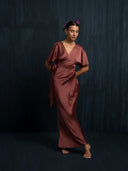 A woman stands barefoot against a dark background, wearing the Ria Silk Dress in burgundy by Rimmba | Sustainable Clothing, hands behind her back and purple flowers in her hair.