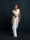Barefoot woman wearing a white blouse cowl neckline and white silk sarong with bronze brosche against a dark background.