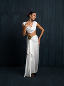 Barefoot woman wearing a white silk linen blouse , cowl neckline and white silk sarong with broze brosche against a dark background.