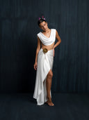 Barefoot woman wearing a white silk linen blouse cowl neckline and white silk sarong with bronze brosche against a dark background.