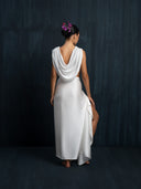 Barefoot woman wearing a white silk linen blouse cowl neckline and white silk sarong is facing a dark background.