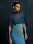 a woman standing against a dark background, wearing an elegant Mulberry silk dress ombre. She has an indigo scraf draped over her shoulders, adding a soft and graceful touch to her look. Her hair is neatly tied back and adorned with purple flowers, giving the outfit a natural and sophisticated appearance.