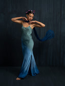 A woman barefoot poses gracefully against a dark background, wearing the Scarlette Silk Dress - Indigo Ombre by Rimmba | Sustainable Clothing, with a blue scarf flowing behind her and small decorative flowers in her hair.