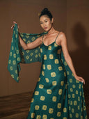 A woman poses indoors in the Scarlette Silk Dress - Emerald Shibori by Rimmba | Sustainable Clothing, featuring yellow square patterns and a matching shawl, standing on a wooden floor with a brown background.