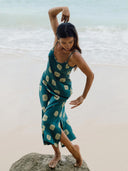 A woman in the Scarlette Silk Dress - Emerald Shibori by Rimmba | Sustainable Clothing stands barefoot on a beach rock, posing gracefully with raised arms, as ocean waves shimmer in the background.