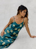 A woman in Rimmba’s Scarlette Silk Dress – Emerald Shibori sits on a light surface, leaning back with long hair and gold earrings, exuding effortless elegance.