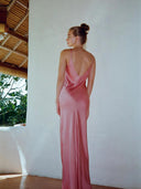 A woman with her hair in a bun stands with her back to the camera, wearing the Rimmba Scarlette Silk Dress - Full Length Maxi in a bright room with white walls and a bamboo ceiling.