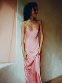 A woman with curly hair smiles indoors, wearing the Scarlette Silk Dress - Full Length Maxi by Rimmba | Sustainable Clothing in pink mulberry silk satin, gazing to the side with her hand near her knee against pastel-lit walls.