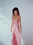 A woman with curly hair stands against a white background, wearing the Scarlette Silk Dress - Full Length Maxi in pink by Rimmba | Sustainable Clothing. She looks down and gently holds the side of her dress.