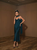 A woman poses confidently on a wooden floor in a minimalist brown-walled room, wearing Rimmba’s Scarlette Silk Dress in indigo and black heels, with one hand on her hip and the other touching her scarf.