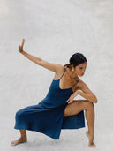 A woman wearing the Scarlette Silk Dress - Indigo by Rimmba | Sustainable Clothing crouches barefoot on sand, one arm extended upward and the other bent, striking a graceful pose that evokes dance or martial arts.