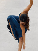 A woman with long brown hair wears the Scarlette Silk Dress - Indigo by Rimmba | Sustainable Clothing, gracefully bending forward with one arm extended and the other raised as if dancing on a light gray surface.