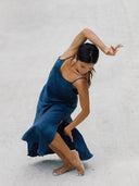 A woman dances barefoot on a light surface in the Scarlette Silk Dress - Indigo by Rimmba | Sustainable Clothing, gracefully bending her knees, raising one arm above her head while the other hand touches her leg.