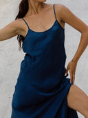 A woman models the Scarlette Silk Dress in Indigo by Rimmba | Sustainable Clothing, raising one arm and holding the high-slit dress at her thigh against a light, neutral background.