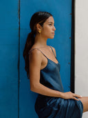 A woman with long brown hair in a ponytail, wearing Rimmba’s Scarlette Silk Dress in indigo, sits sideways against a vibrant blue door, gazing ahead with a calm expression.