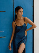 A woman wearing the Scarlette Silk Dress in Indigo by Rimmba | Sustainable Clothing stands by a bright blue door, looking to the side. Her long dark hair is pulled back and she accessorizes with gold hoop earrings.