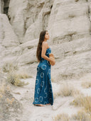 A woman in the Scarlette Silk Dress - Indigo Circle by Rimmba | Sustainable Clothing stands barefoot on sandy, rocky ground with light rock formations behind her, holding a tan bag as the dress’s white squares and indigo circles flow around her.