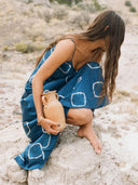 A woman in the Scarlette Silk Dress - Indigo Circle by Rimmba | Sustainable Clothing crouches barefoot on rocks, holding a clay jug amid dry grass and shrubs, her long hair partly obscuring her face.