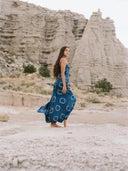 A woman in the Scarlette Silk Dress - Indigo Circle by Rimmba | Sustainable Clothing stands barefoot on sand before rocky formations, her long dark hair flowing as she gazes calmly into the distance.