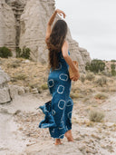A woman with long brown hair wears the Scarlette Silk Dress - Indigo Circle by Rimmba | Sustainable Clothing, standing barefoot on rocky ground with one arm raised amid a desert landscape and dramatic rock formations.
