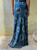 A person stands on stone tiles wearing the Rimmba | Sustainable Clothing Scarlette Silk Dress - Indigo Circle, its deep blue silk and circular patterns draping elegantly against a simple wall, viewed from behind.